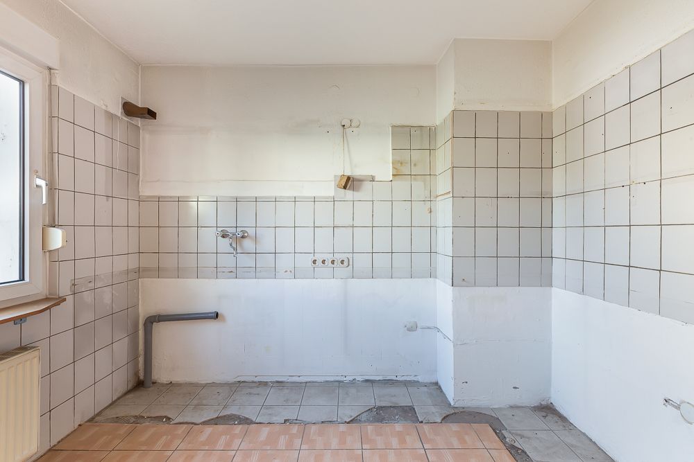 Leerer Küchenraum mit weißen Fliesen und sichtbaren Wasseranschlüssen.