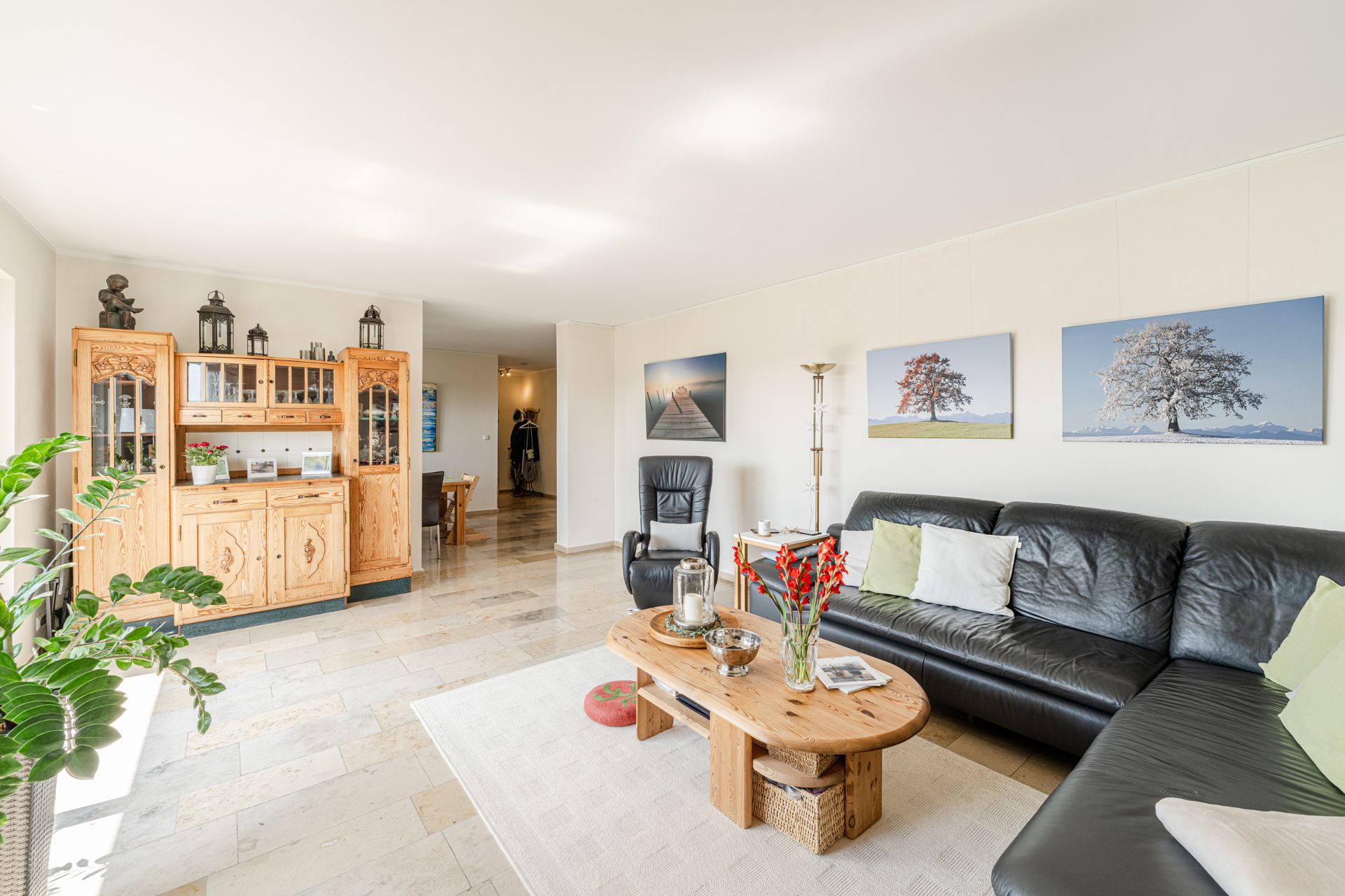Helles Wohnzimmer mit schwarzem Ledersofa, Holztisch, Vitrine und Wandbildern von Bäumen.