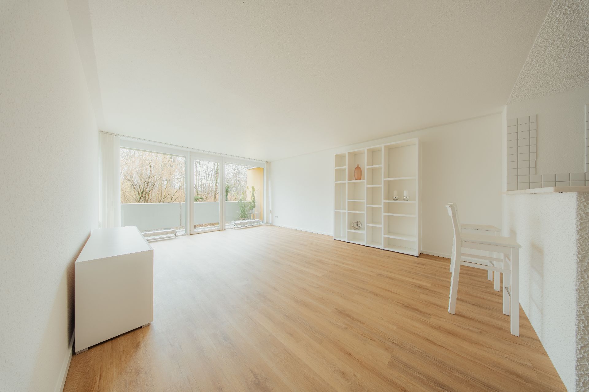 Helles Wohnzimmer mit Holzboden, großen Fenstern, Regal und kleinem Esstisch mit zwei Stühlen.
