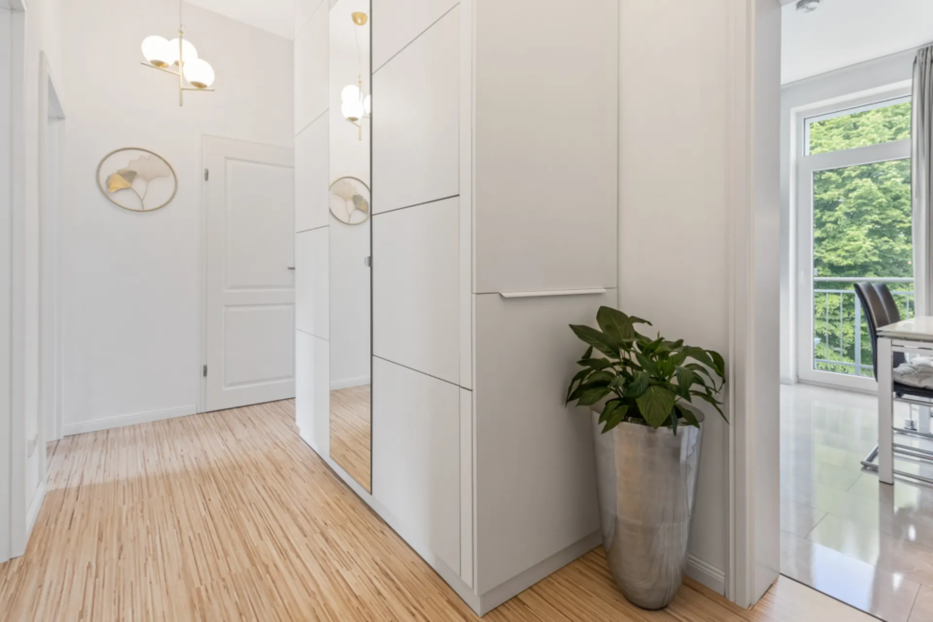 Heller Flur mit Holzfußboden, weißem Schrank mit Spiegel und großer Pflanze im Metalltopf.