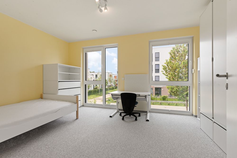 Helles Zimmer mit Einzelbett, Schreibtisch, Stuhl, Regal und großem Fenster.