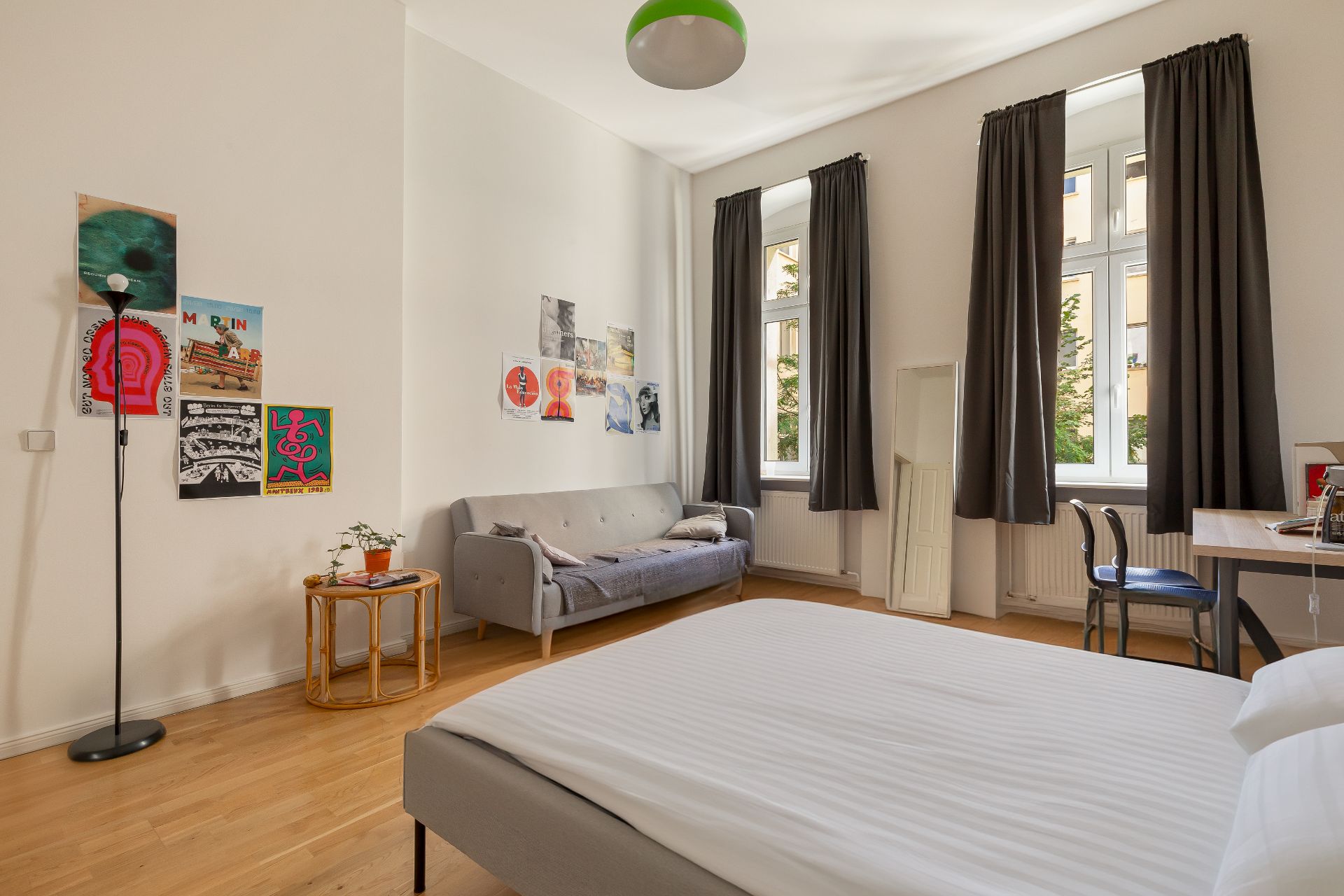 Helles Schlafzimmer mit Bett, Sofa, Schreibtisch, drei Fenstern und Poster an der Wand.