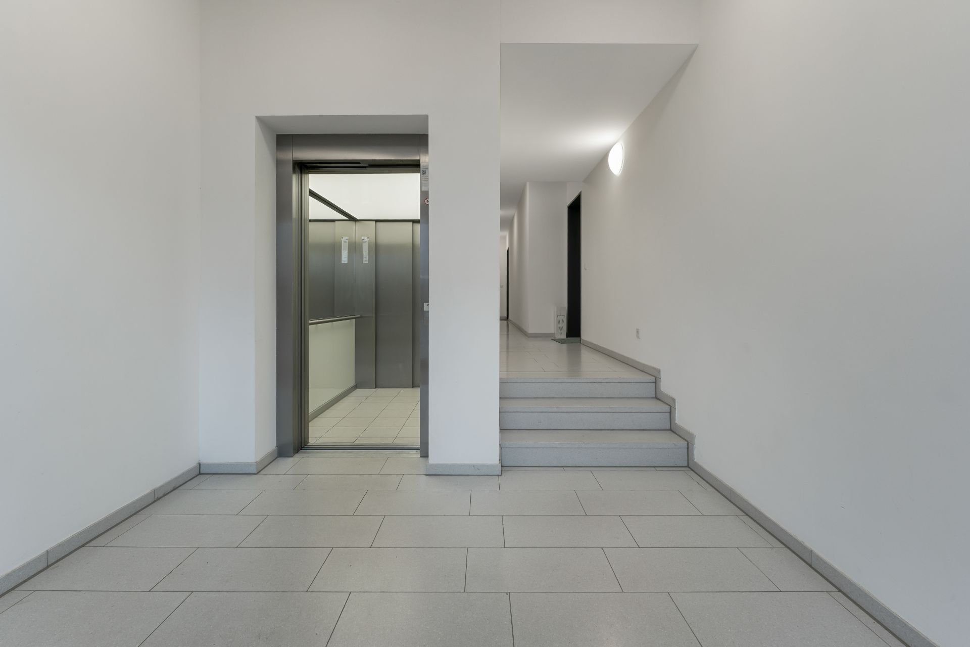 Heller Flur mit Aufzug und einigen Treppen, weiße Wände und graue Fliesen.