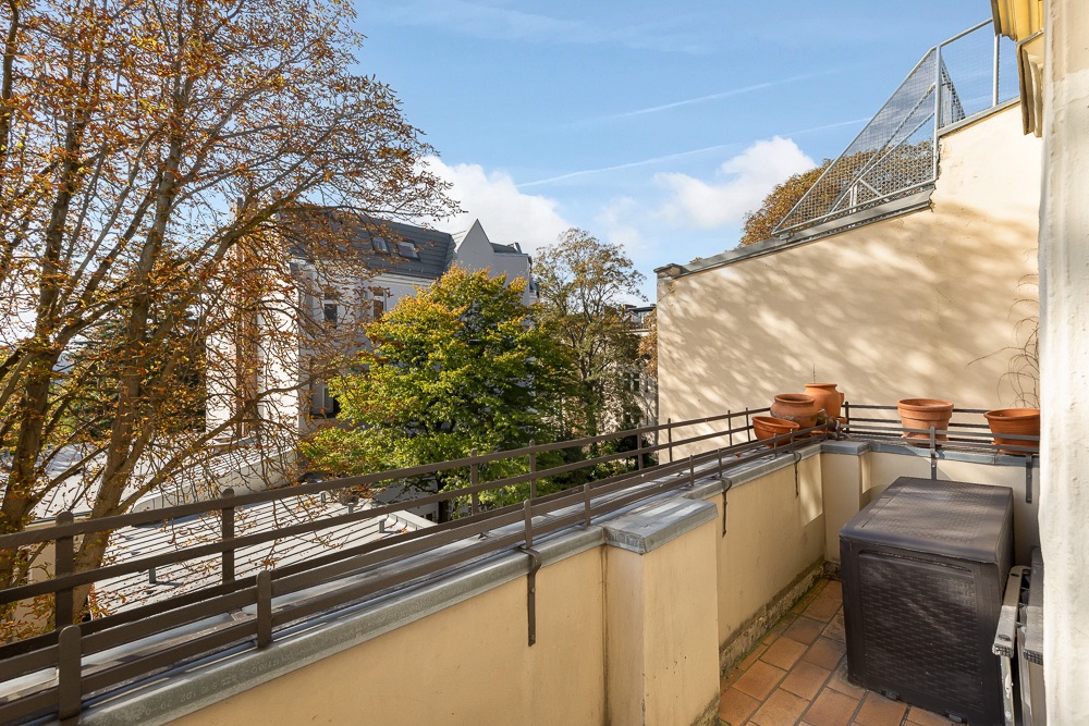 Balkon mit Geländer, mehreren Pflanztöpfen und Blick auf Bäume sowie Nachbargebäude.