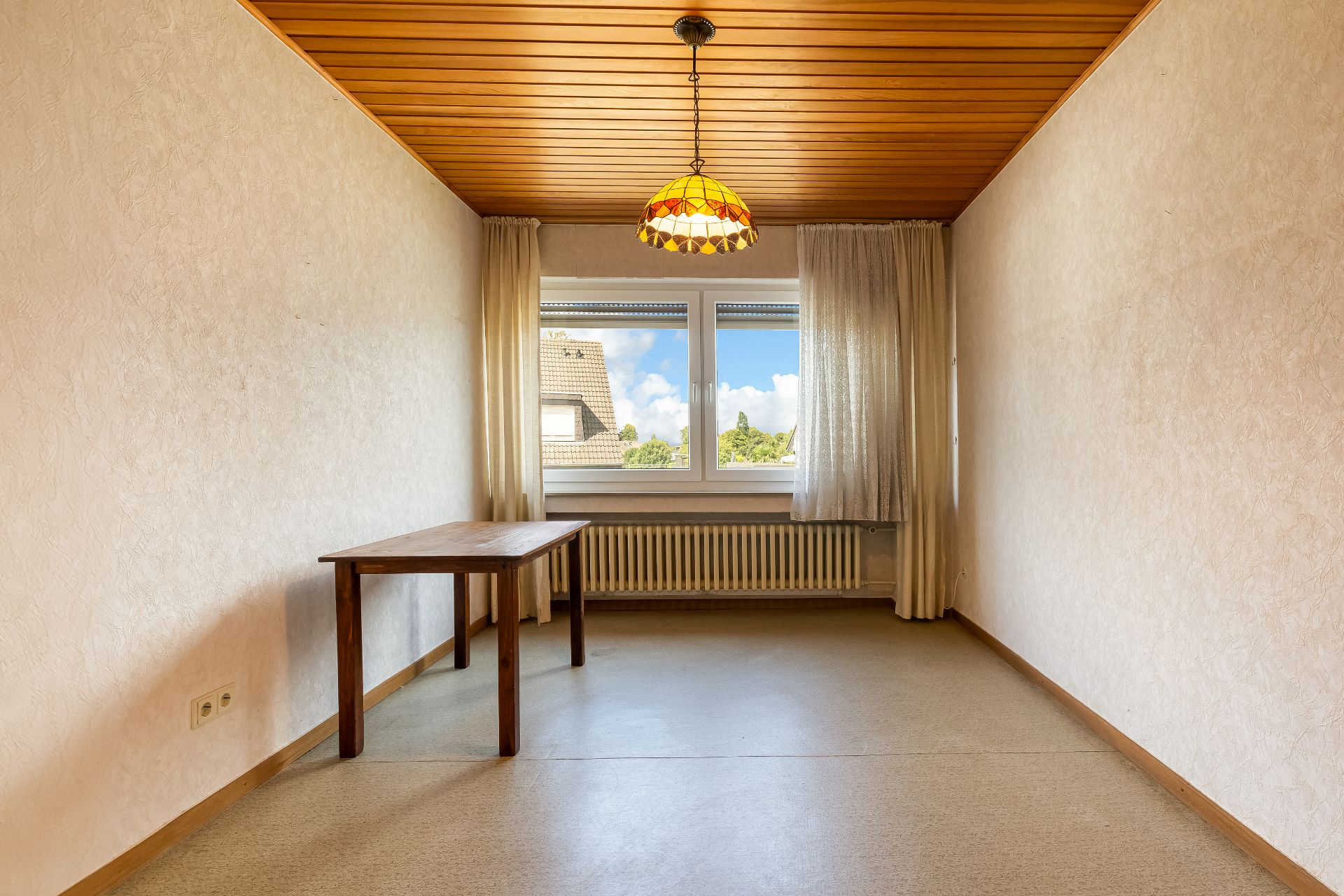 Schmaler Raum mit Holztisch, Fenster mit Vorhängen und Deckenlampe.