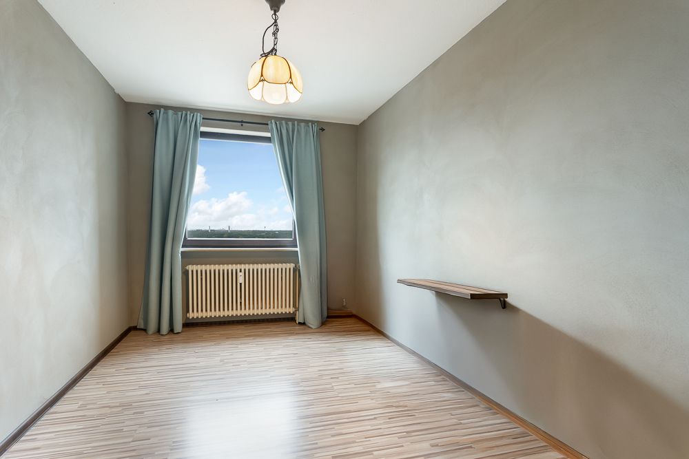 Leeres Zimmer mit Holzfußboden, Fenster mit Vorhängen, Heizung und einer kleinen Wandablage.