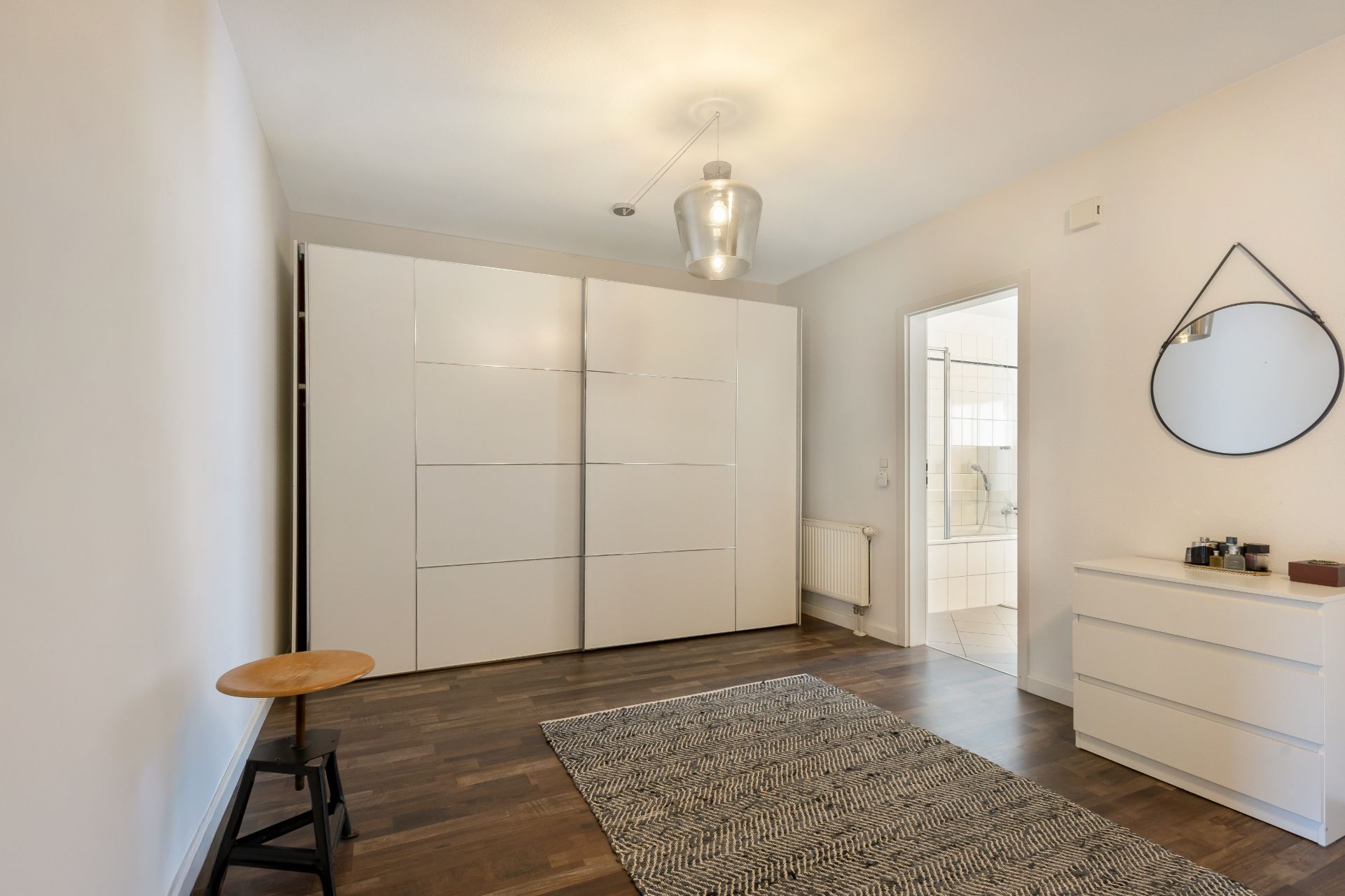 Helles Schlafzimmer mit weißem Schrank, Kommode, Spiegel und Teppich auf dunklem Holzboden.