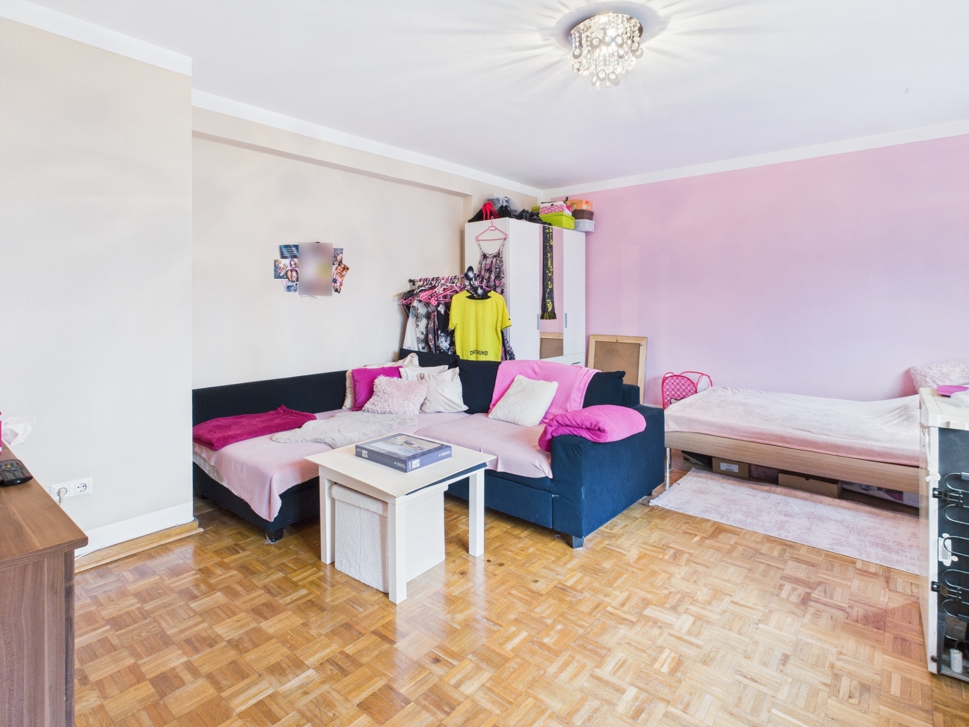 Helles Zimmer mit Sofa, Bett, Couchtisch und Kleiderschrank auf Parkettboden.