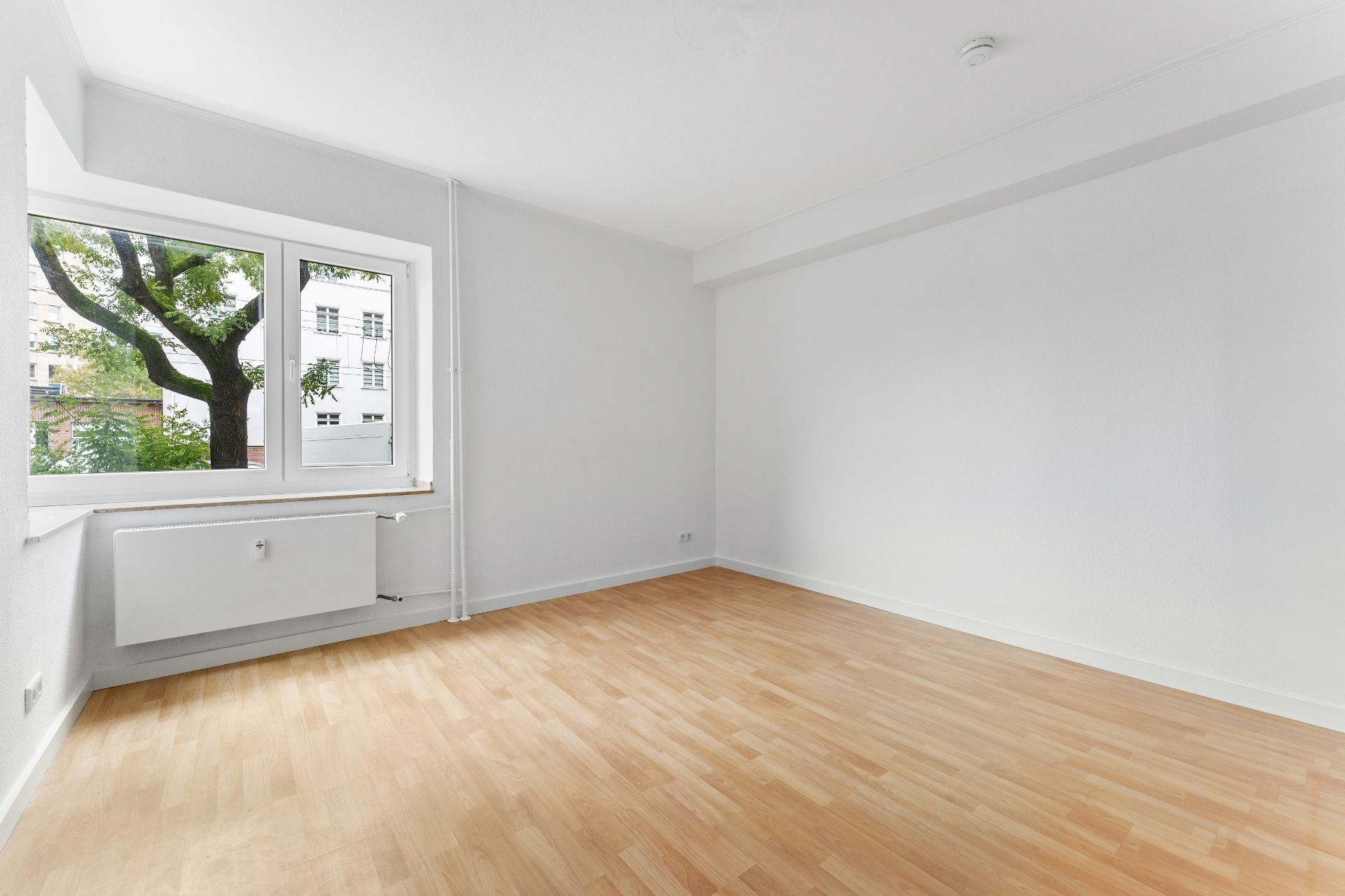 Helles, leeres Zimmer mit Holzfußboden, weißer Wand und großem Fenster.