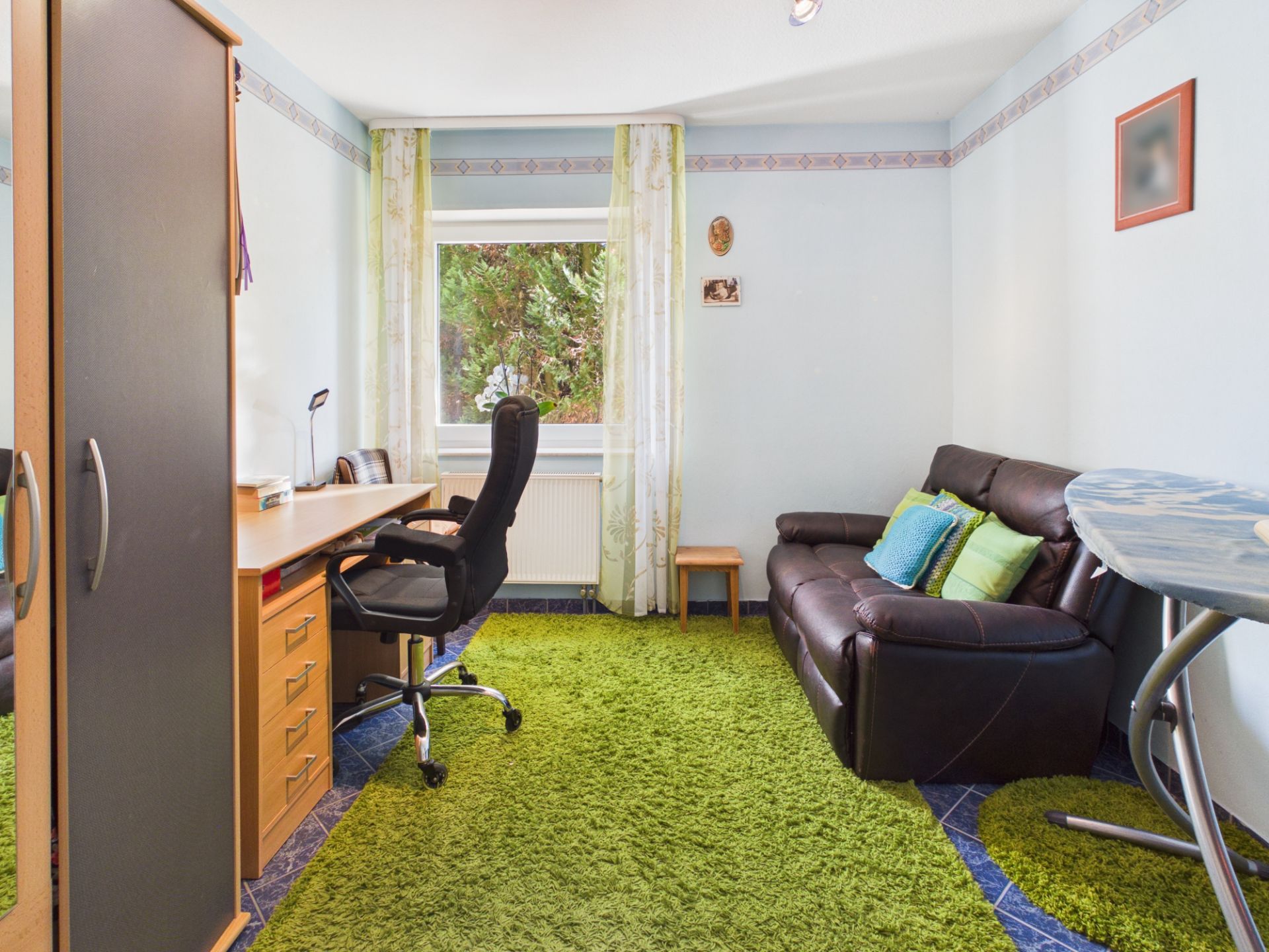 Helles Zimmer mit Schreibtisch, Drehstuhl, brauner Couch und grünem Teppich.