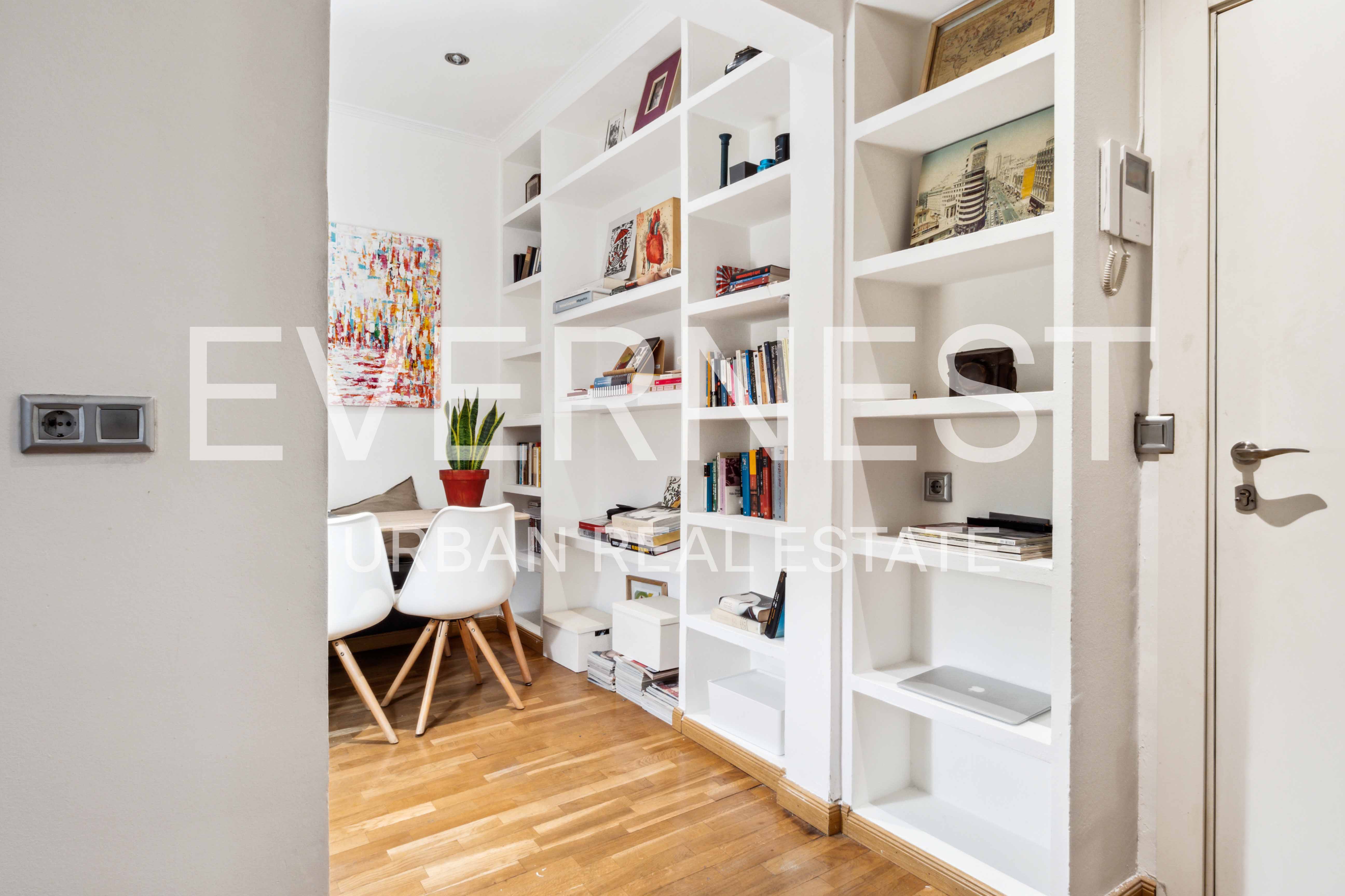 Helles Zimmer mit weißen Regalen, Holzboden, zwei weißen Stühlen, Tisch und bunter Wandkunst.