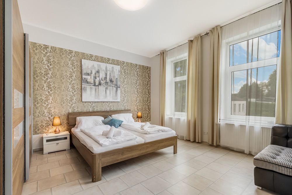Helles Schlafzimmer mit großem Bett, zwei Nachttischen, Fenstern mit Vorhängen und gemusterten Tapeten.