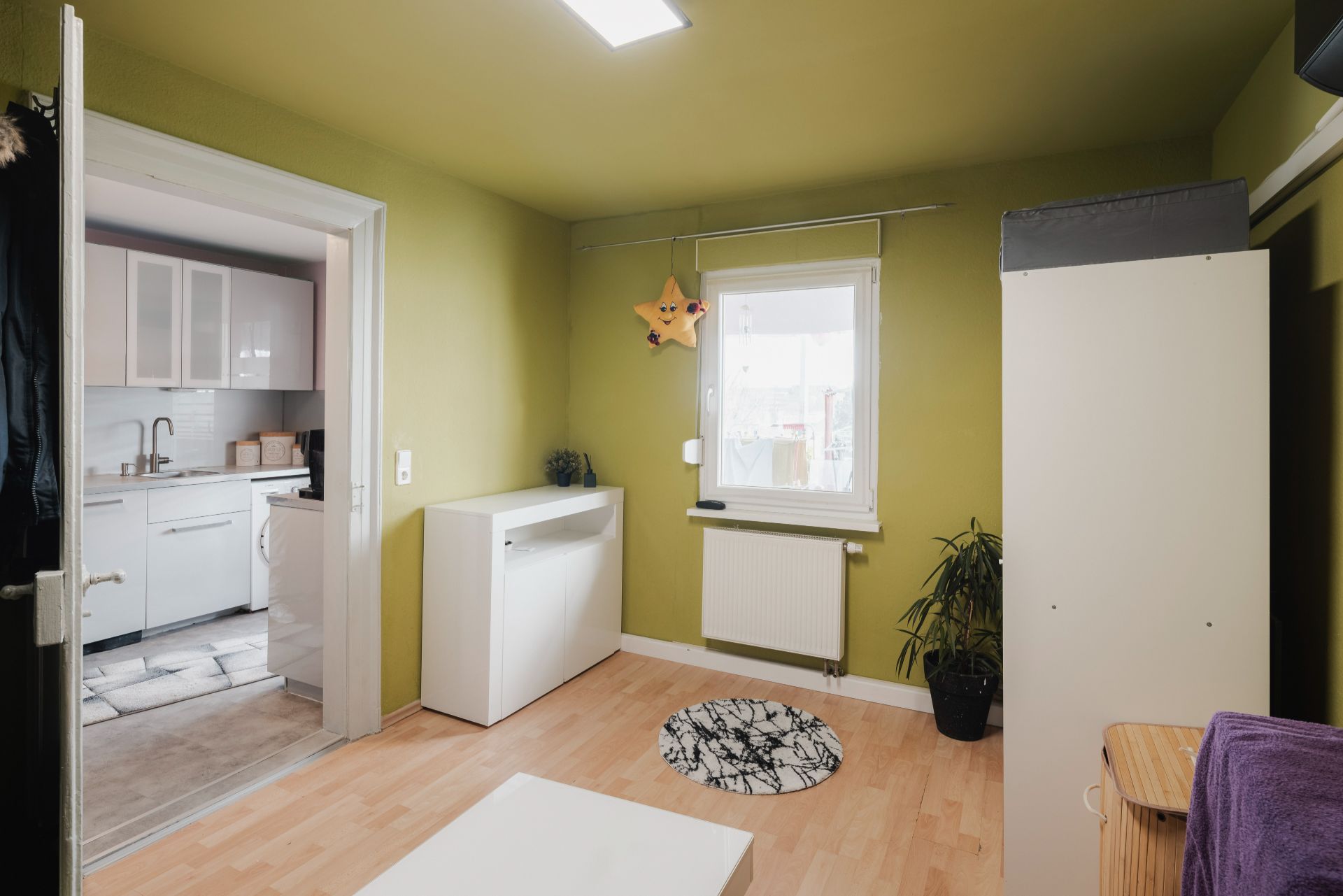 Kleines Zimmer mit grün gestrichenen Wänden, Fenster, Pflanze und Tür zur weißen Küche.