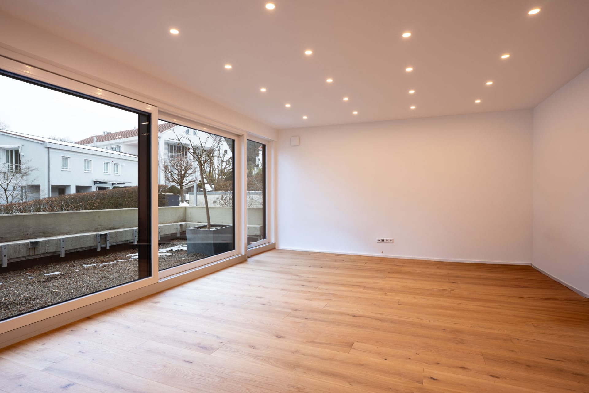 Leerer, heller Raum mit Holzfußboden und großen Fenstern zur Terrasse.