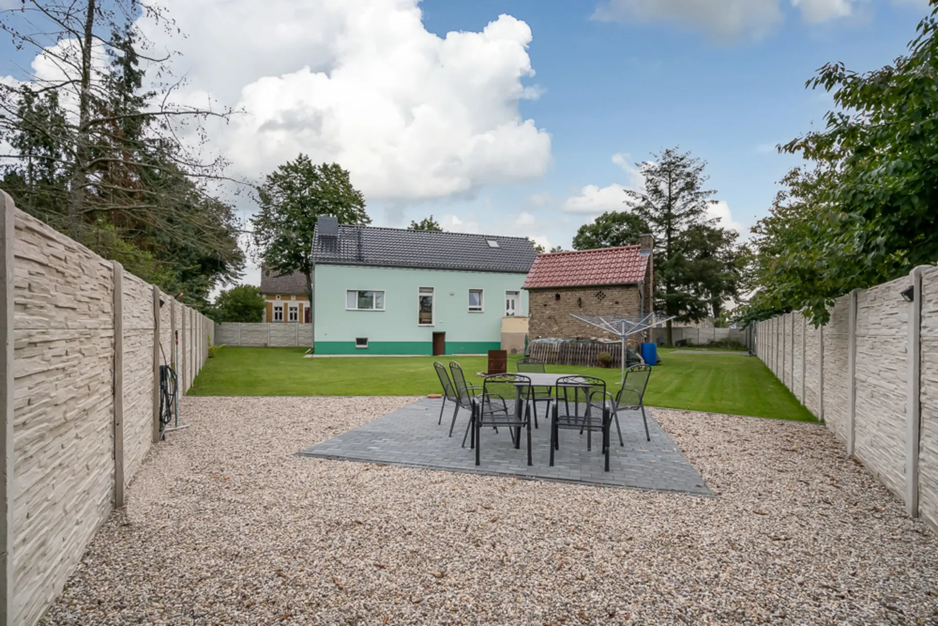 Garten mit Sitzgruppe auf Steinboden, Wäscheleine und grünem Rasen, Haus im Hintergrund.
