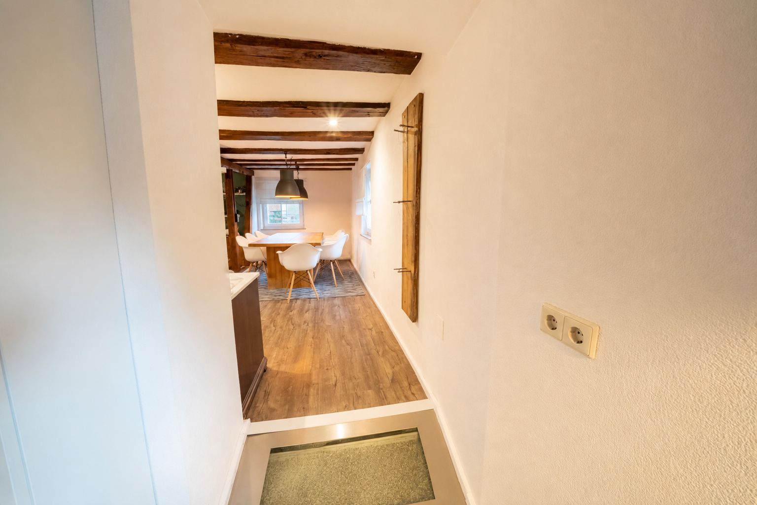 Flur mit Blick in ein Esszimmer mit Holzboden, Holzbalken und Tisch mit weißen Stühlen.