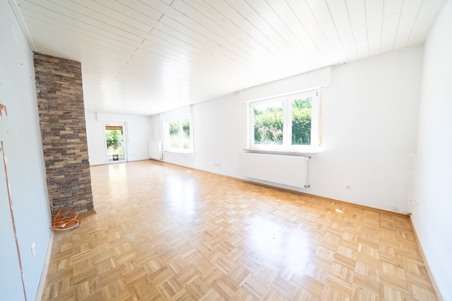 Helles, leeres Wohnzimmer mit Parkettboden, weißen Wänden und großen Fenstern.