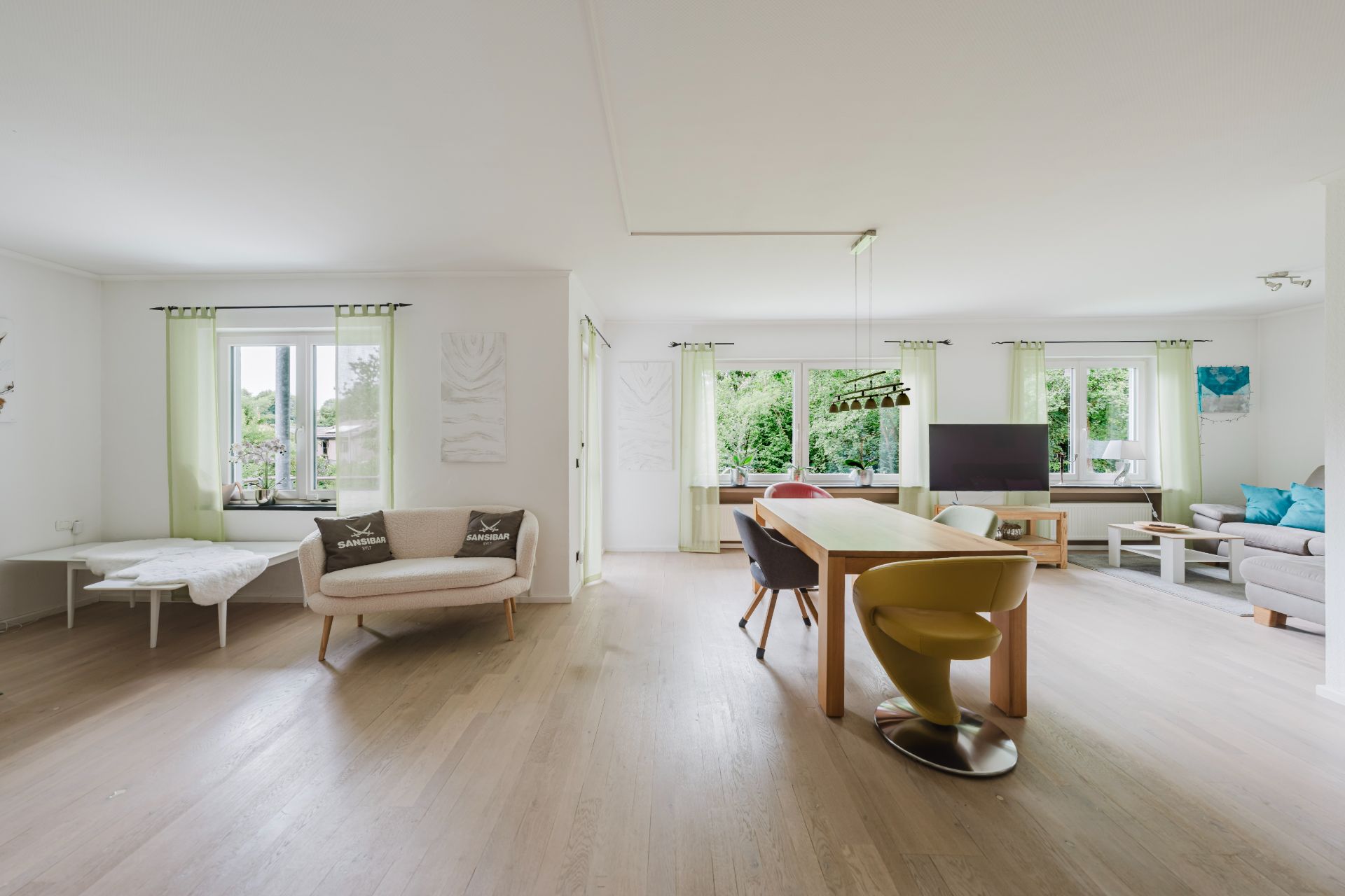 Helles Wohnzimmer mit großem Esstisch, Stühlen, Sofa und großen Fenstern mit Blick ins Grüne.
