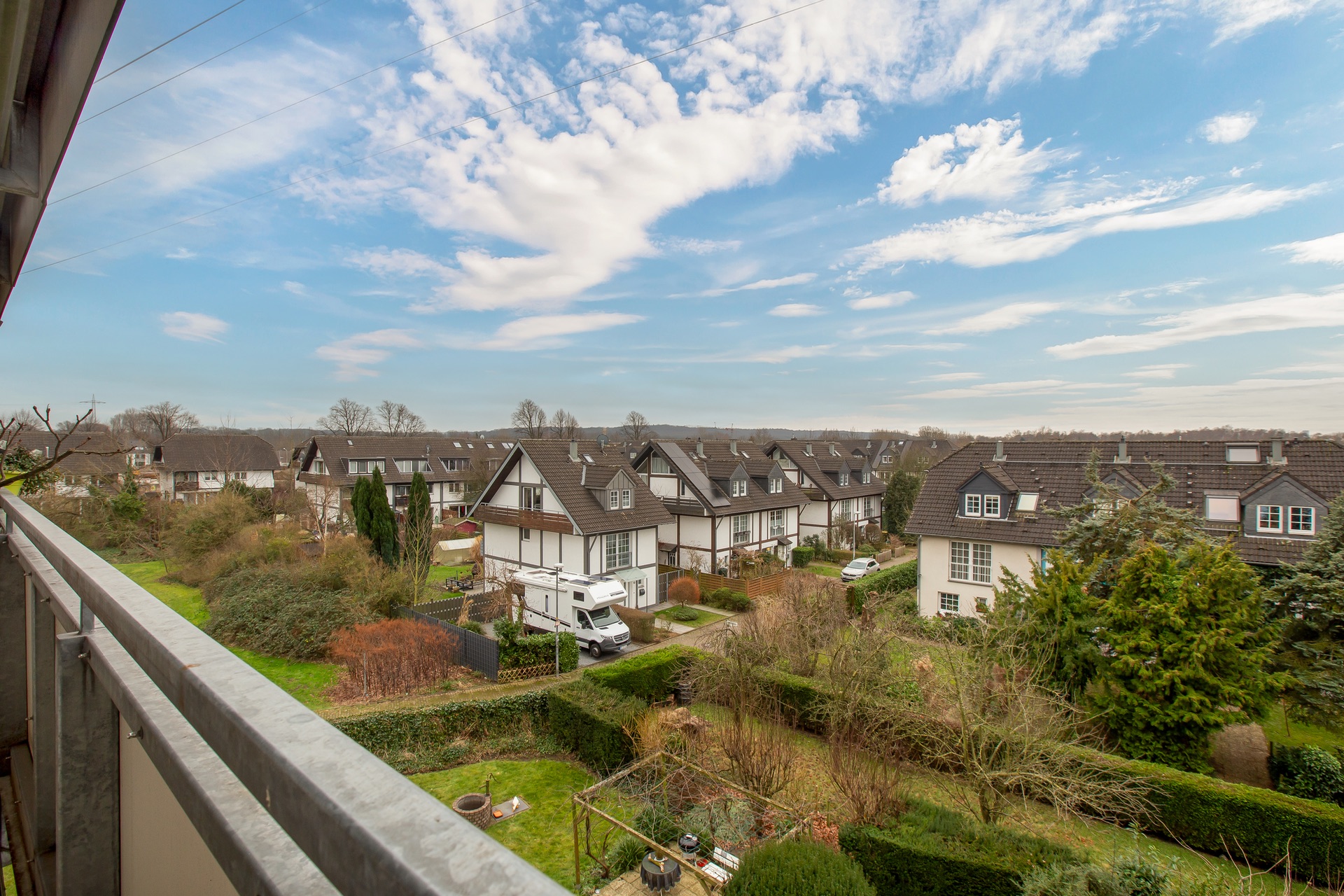 Blick vom Balkon auf Reihenhäuser mit Gärten und Bäumen unter blauem Himmel.
