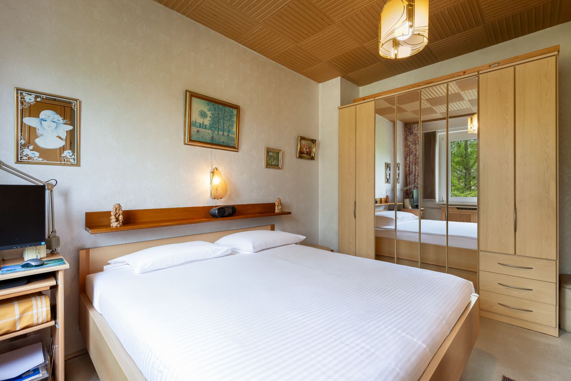Schlafzimmer mit Doppelbett, großem Spiegelschrank und Wandbildern, daneben ein kleiner Schreibtisch mit Fernseher.