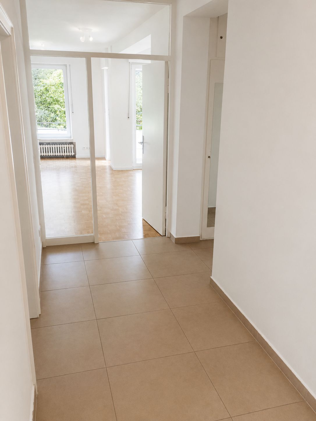 Heller Flur mit Fliesenboden, führt zu einem Raum mit großen Fenstern und Parkett.