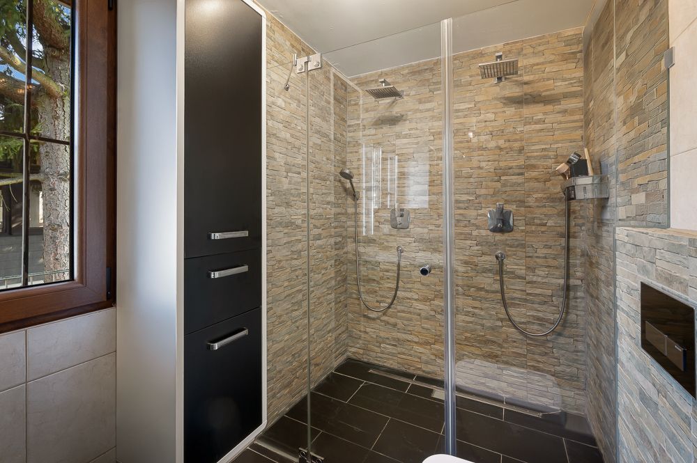 Modernes Badezimmer mit großer, begehbarer Dusche und zwei Duschköpfen, daneben ein Fenster und ein schwarzer Schrank.