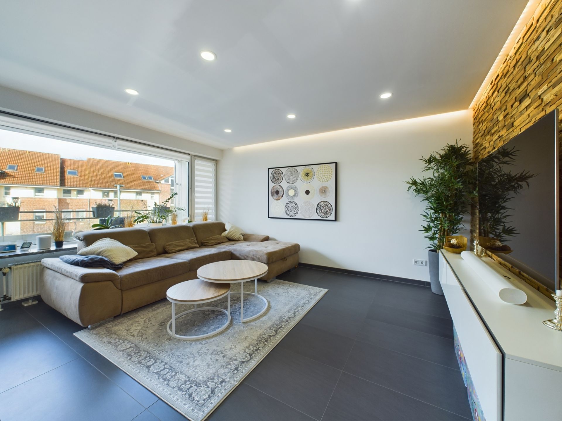 Modernes Wohnzimmer mit großem Sofa, Couchtischen, Teppich und Wanddekoration vor großem Fenster.