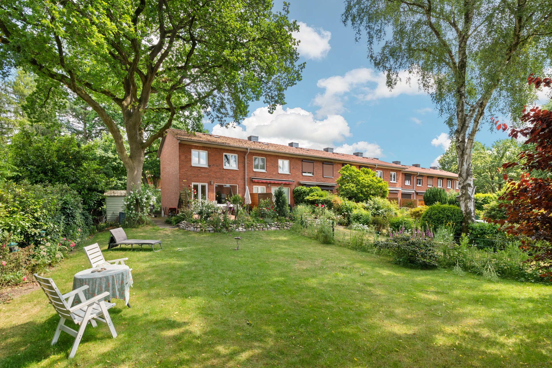 Reihenhaus mit großem Garten, Bäumen, Blumenbeeten und Gartenmöbeln unter freiem Himmel.