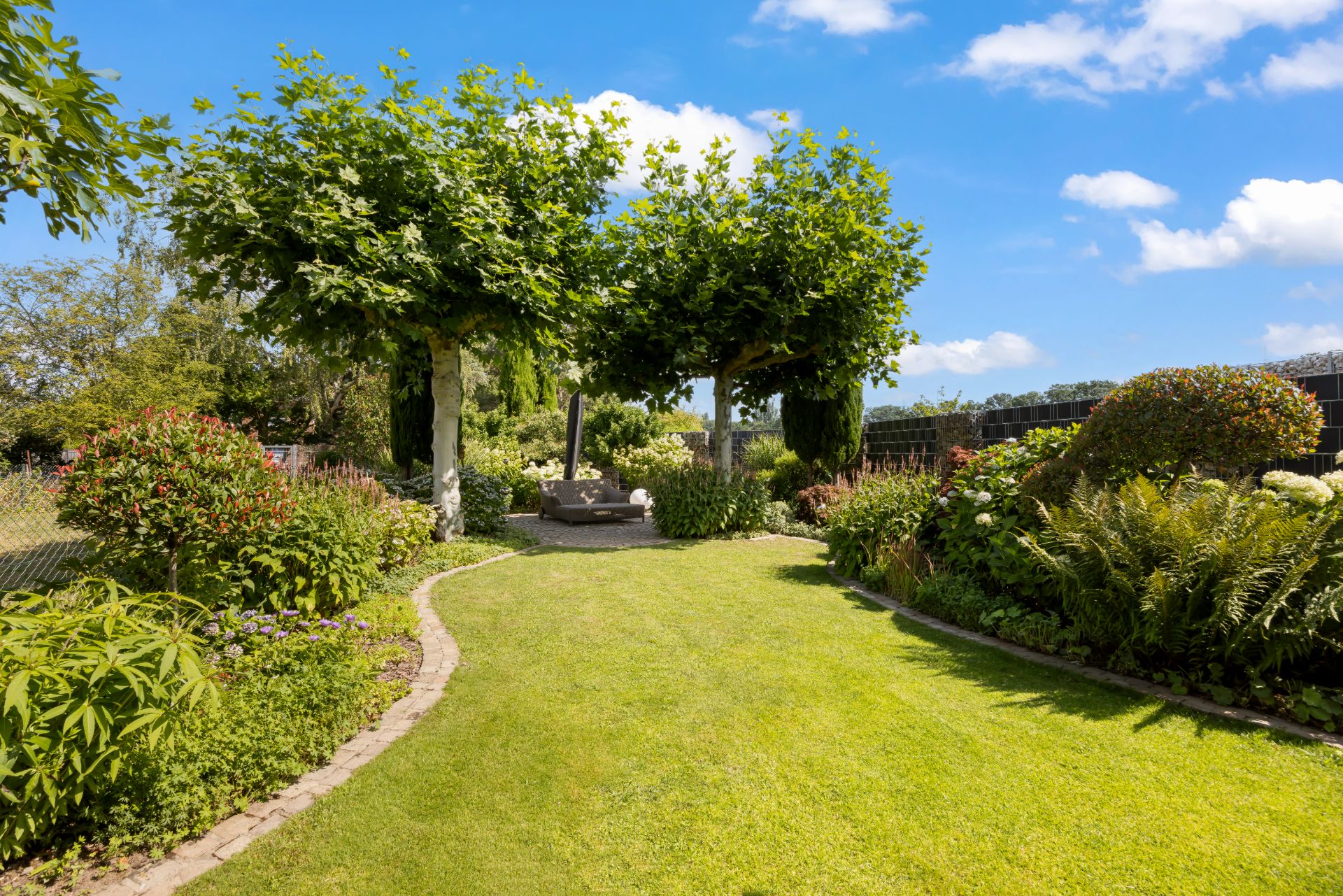 Gepflegter Garten mit Rasen, Bäumen und Blumenbeeten unter blauem Himmel.