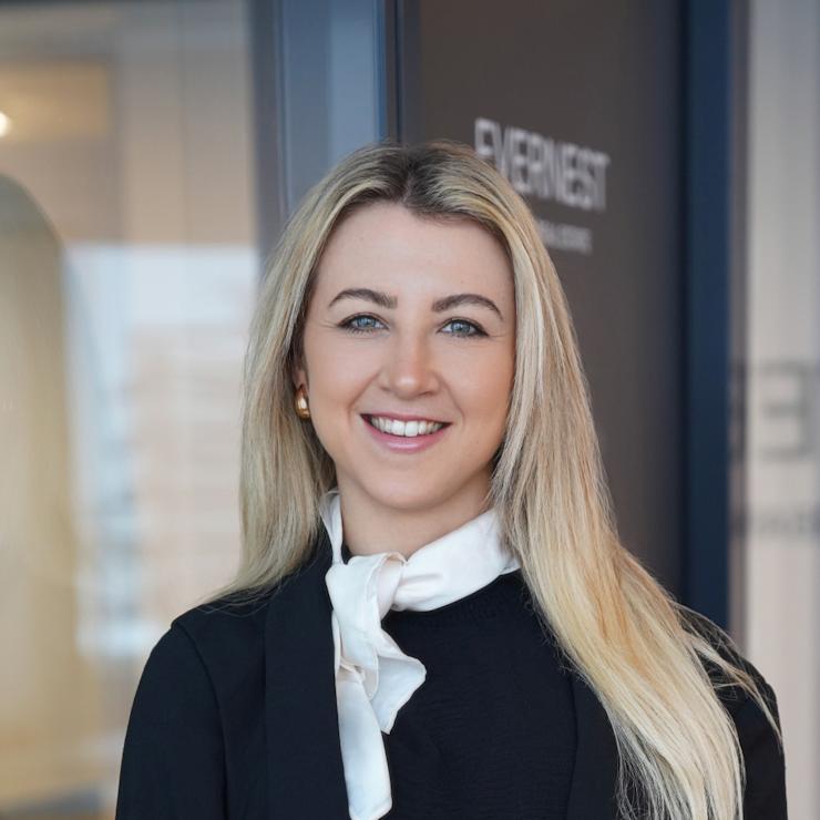 Eine Frau mit langem blondem Haar und schwarzer Kleidung steht und lächelt in einem modernen Innenraum.