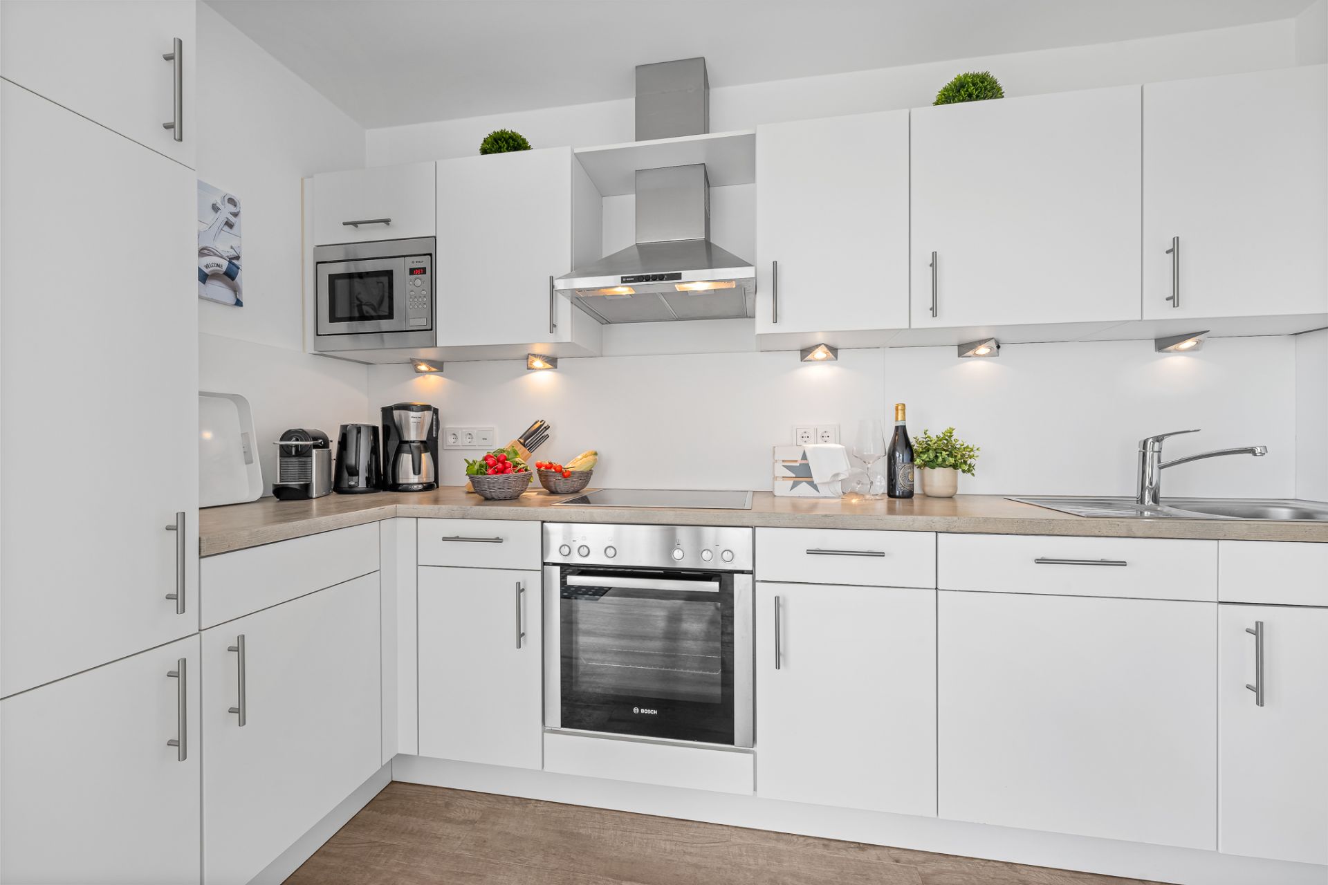 Moderne weiße Einbauküche mit Herd, Spüle, Kaffeemaschine und Obstschale.