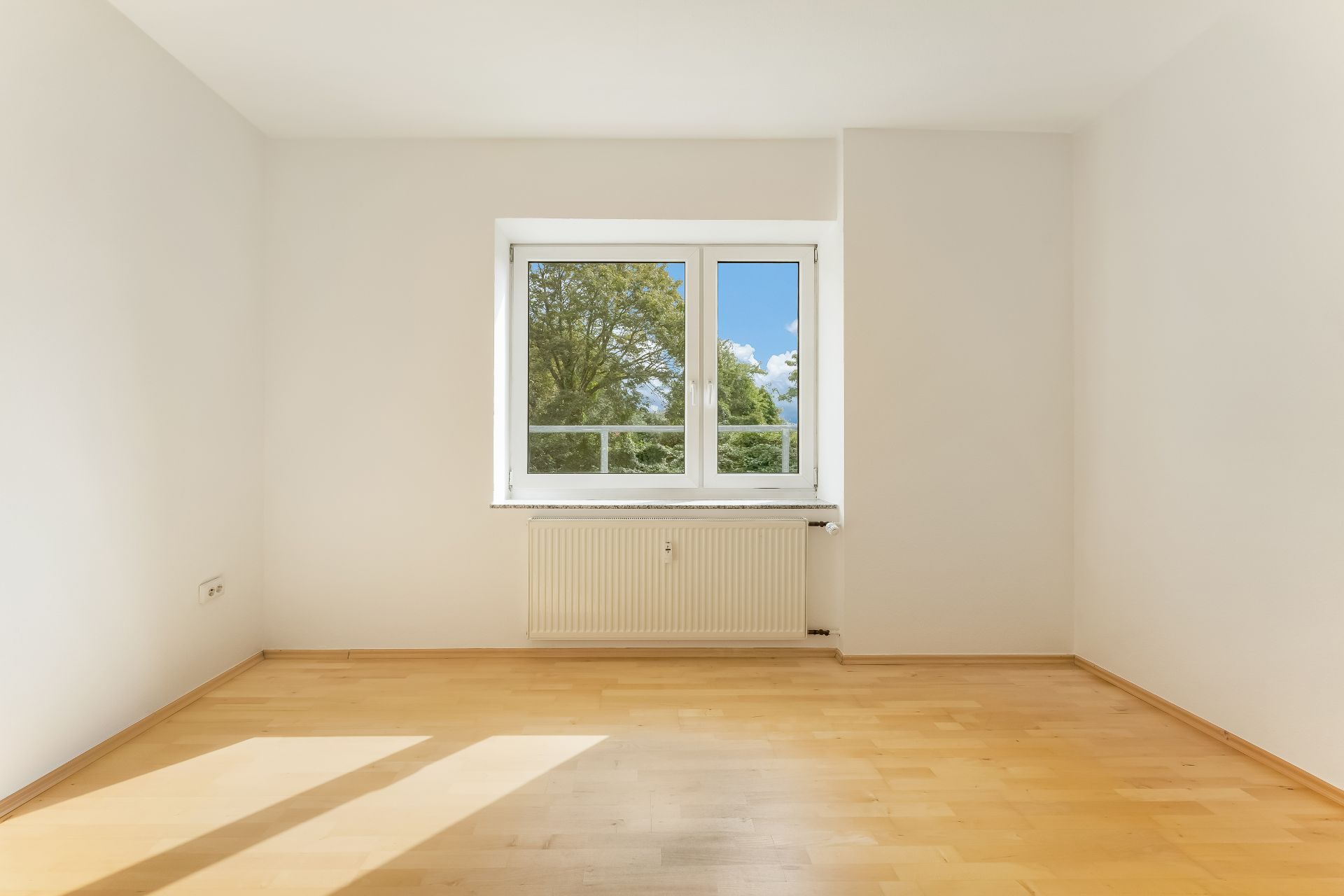 Leerer, heller Raum mit Holzfußboden und großem Fenster, Blick auf Bäume.