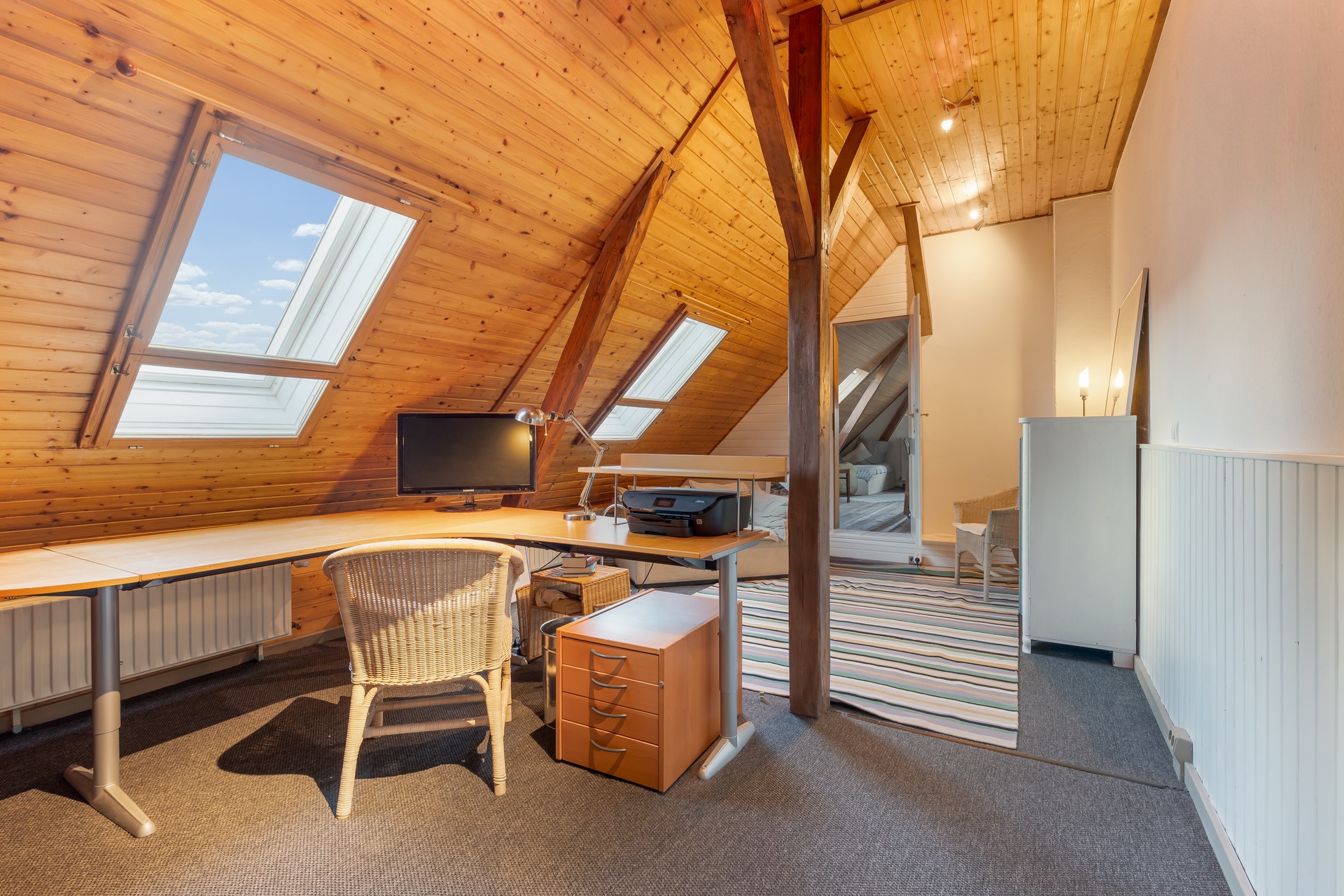 Dachzimmer mit Holzdecke, Schreibtisch, Stuhl, Computer und zwei Dachfenstern.
