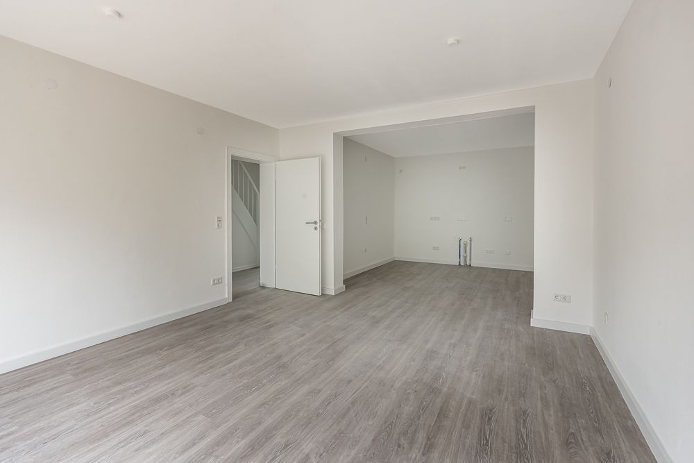 Leerer Raum mit heller Wand, grauem Boden und offener Tür zu einem weiteren Zimmer.