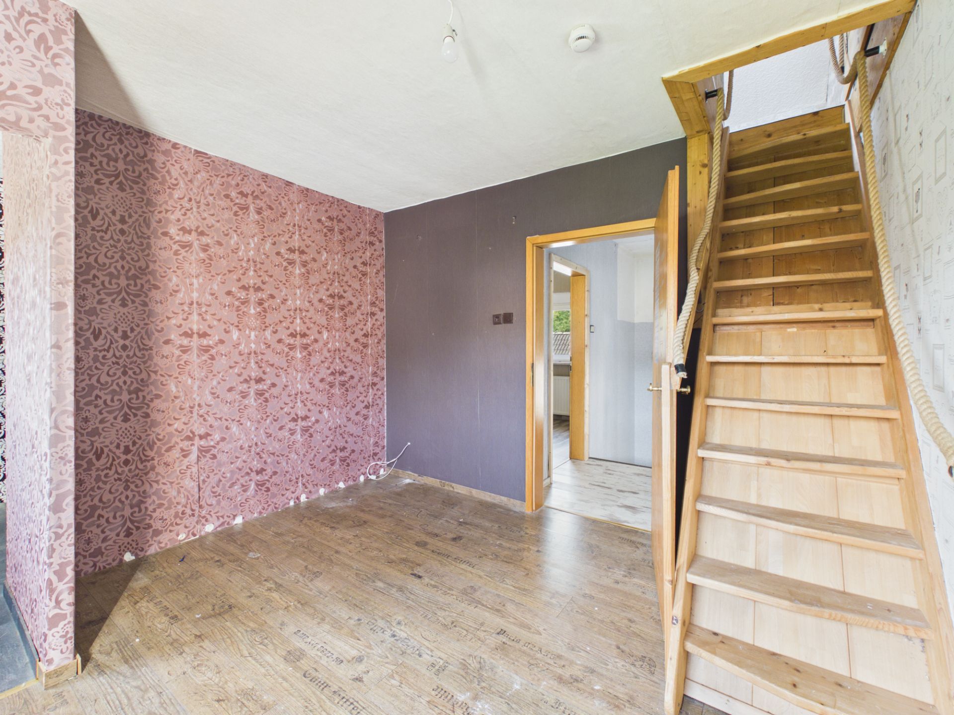 Leeres Zimmer mit gemusterten Tapeten, Holzboden und Holztreppe nach oben.