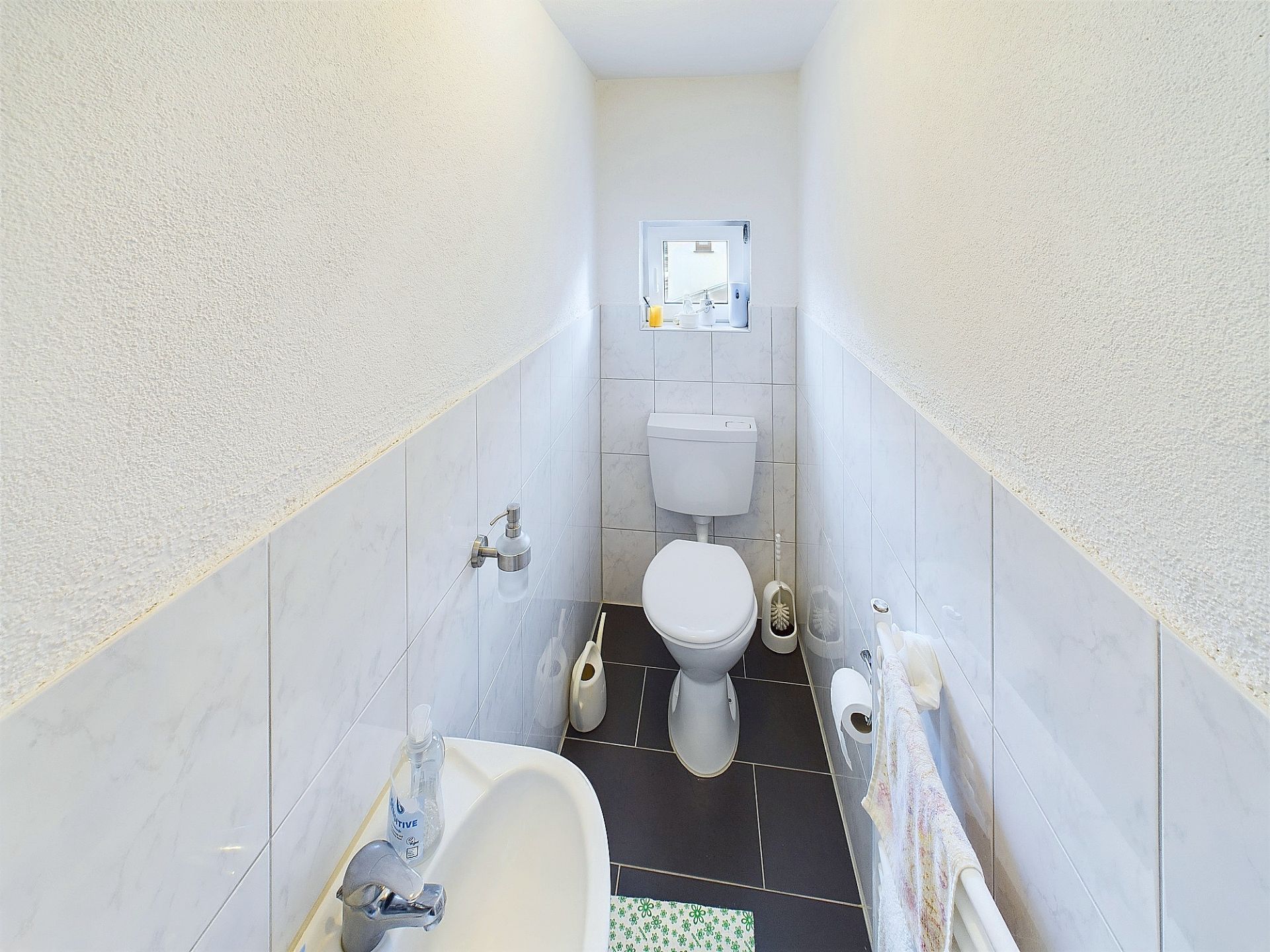 Schmales Badezimmer mit Toilette, Waschbecken, Handtuchhalter und kleinem Fenster.