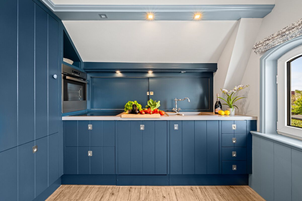 Moderne blaue Einbauküche mit Holzarbeitsplatte, Gemüse, Obst und Dekopflanze am Fenster.