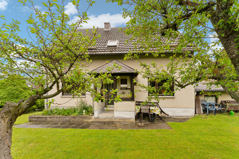 Vorderseite eines Einfamilienhauses mit Garten, Bäumen und Gartenmöbeln auf einer Terrasse.