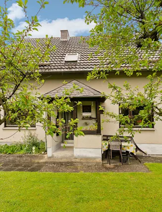 Vorderseite eines Einfamilienhauses mit Garten, Bäumen und Gartenmöbeln auf einer Terrasse.