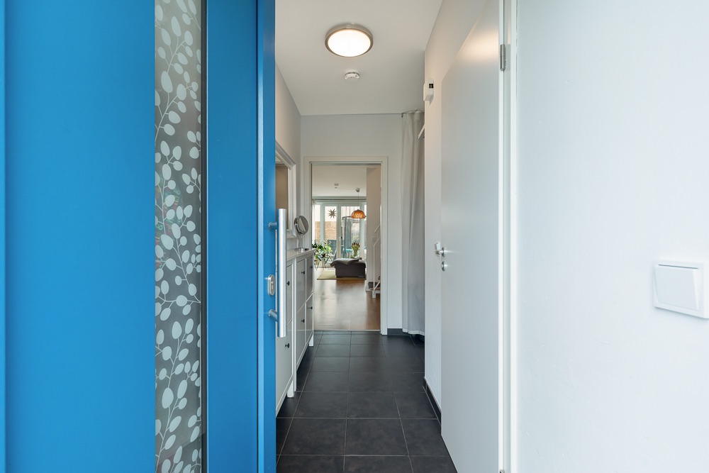 Eingangsflur mit blauer Tür, Fliesenboden und Blick ins Wohnzimmer mit großen Fenstern.