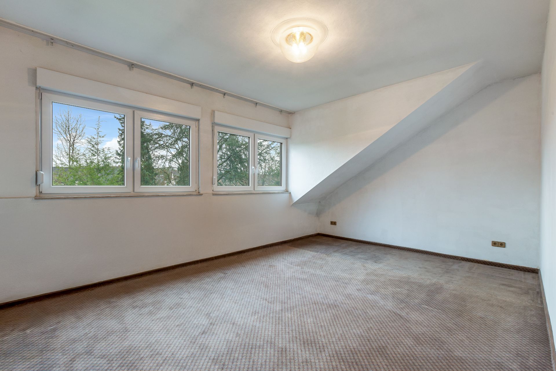 Leeres Zimmer mit braunem Teppich, weißen Wänden und drei Fenstern mit Blick ins Grüne.