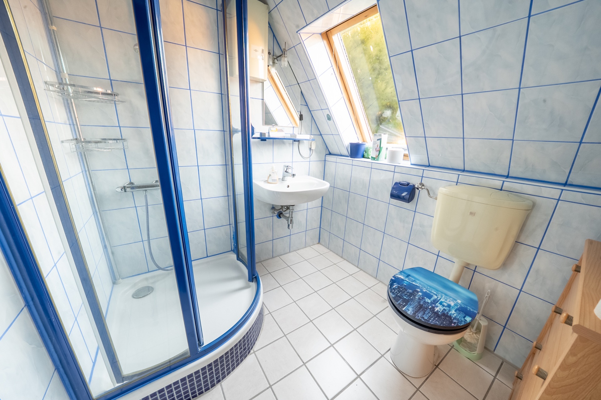 Gefliestes Badezimmer mit Dusche, Waschbecken, WC und zwei Fenstern im Dachbereich.