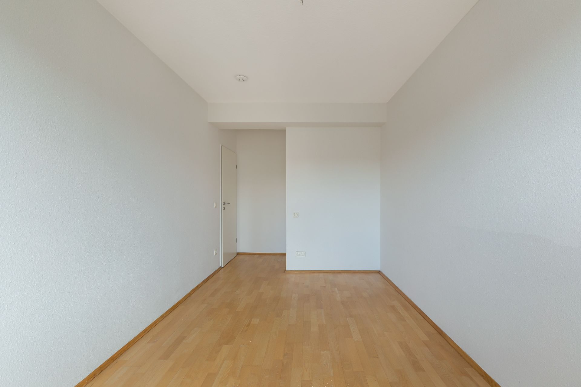 Leerer Raum mit hellem Holzboden und weißen Wänden, Tür im Hintergrund.