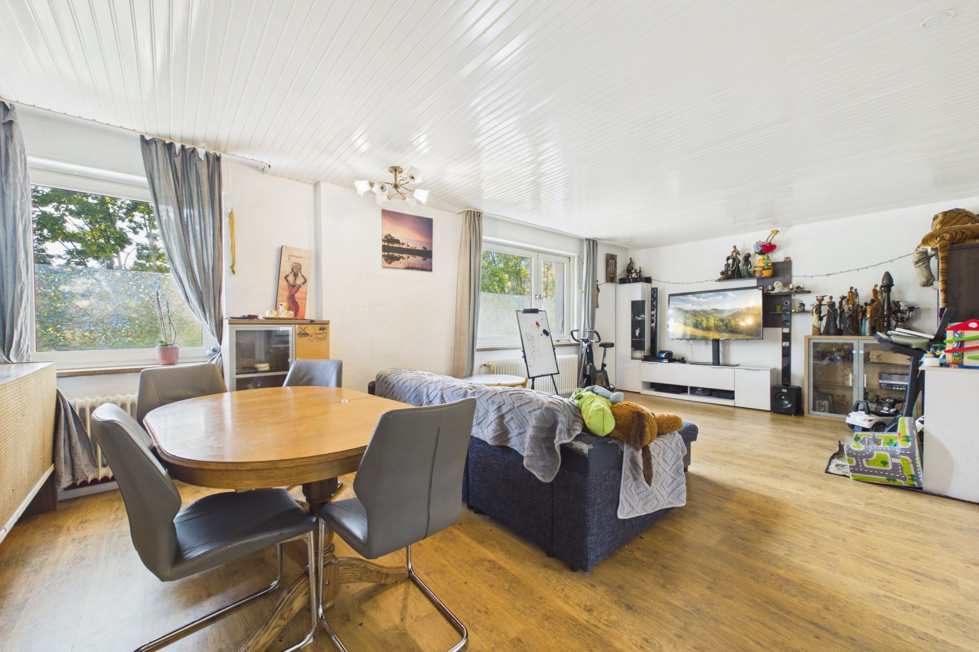 Helles Wohnzimmer mit Essbereich, Sofa, Fernseher und großen Fenstern.