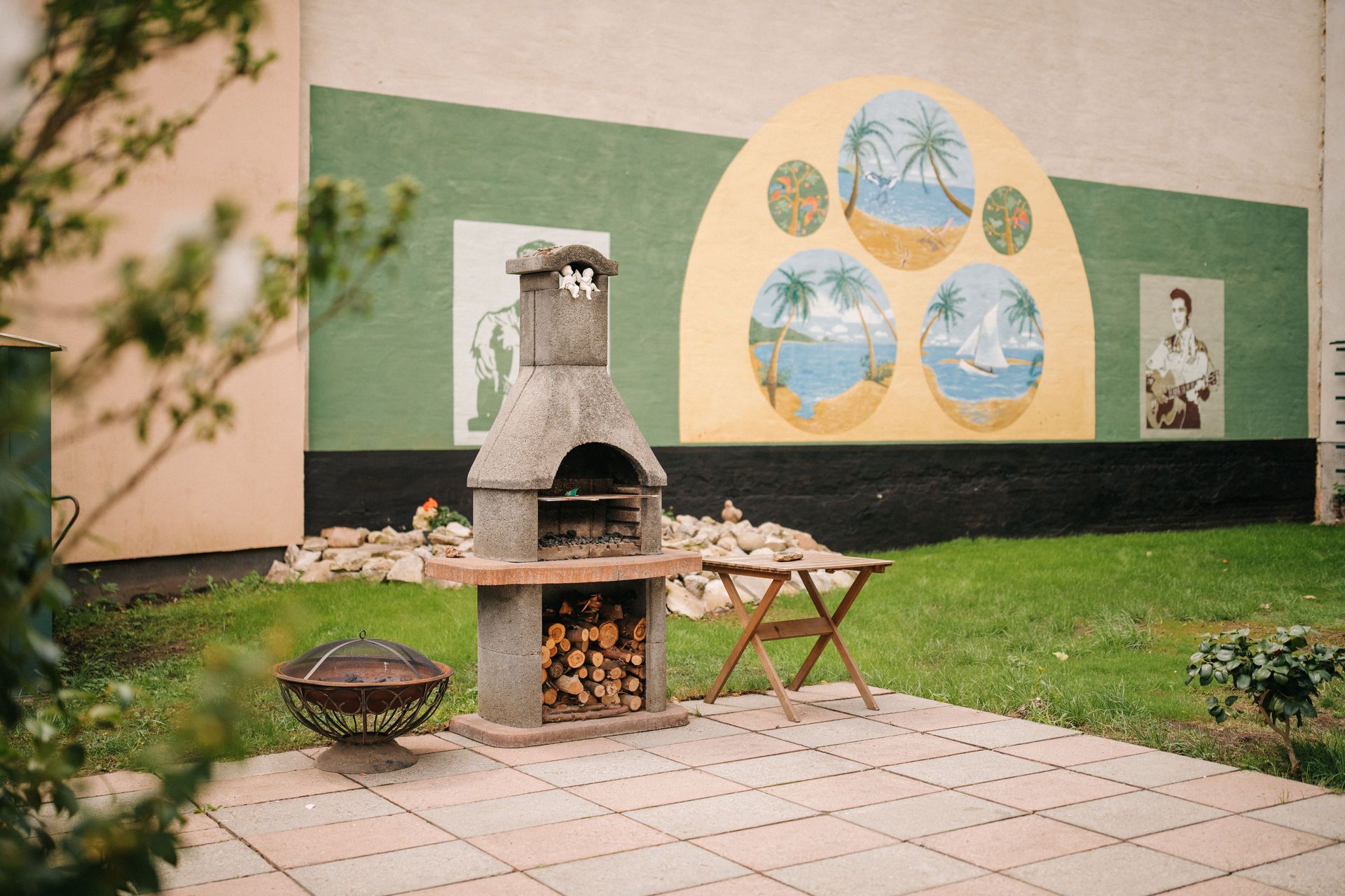 Garten mit festem Grill, kleinem Tisch und Wandbild im Hintergrund.