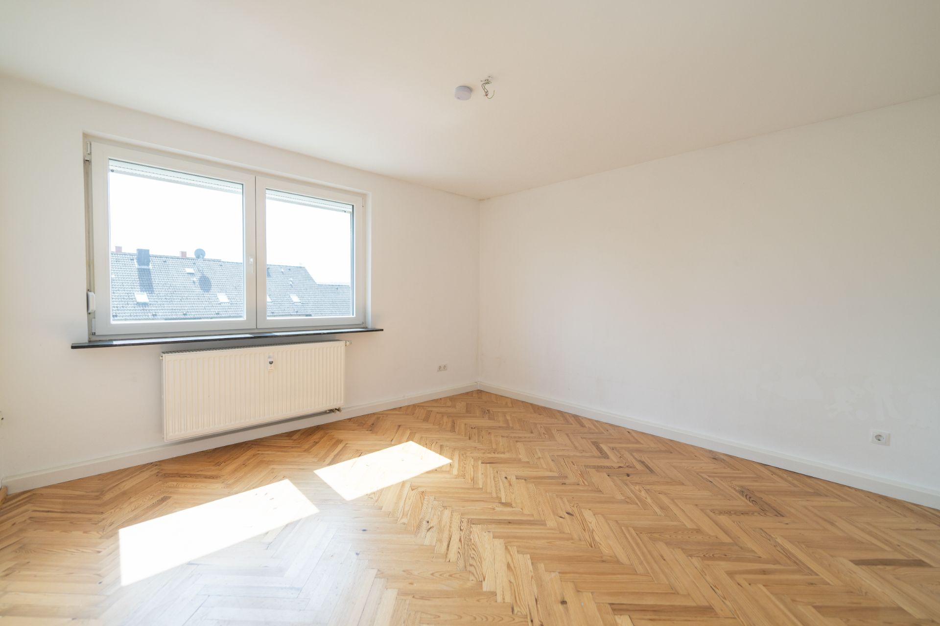 Leeres Zimmer mit Parkettboden, zwei Fenstern und Heizkörper unter dem Fenster.