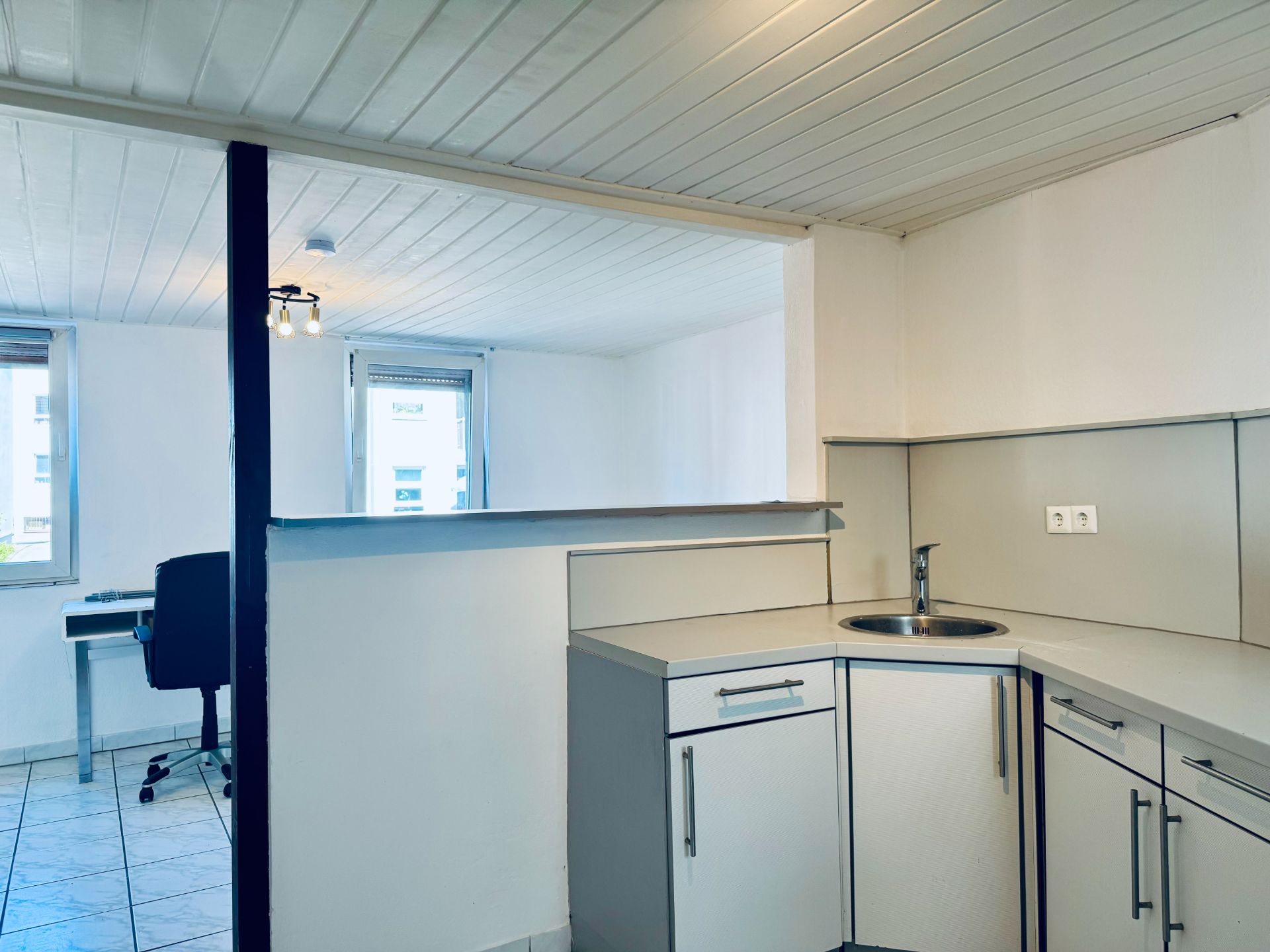 Helle Küche mit Eckspüle, daneben ein Zimmer mit Schreibtisch und Stuhl vor zwei Fenstern.