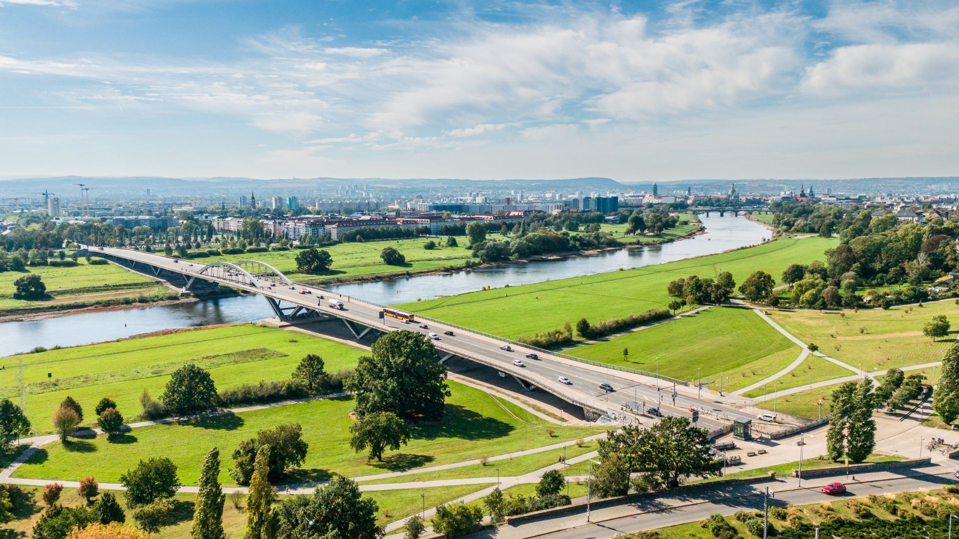 Blick auf eine Brücke über einen Fluss mit viel Grünfläche und Bäumen im Vordergrund.