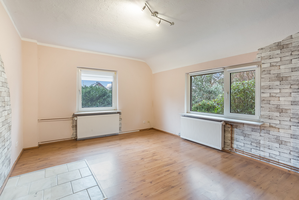 Helles Wohnzimmer mit Holzboden, zwei Fenstern und Heizkörpern, eine Wand teils mit Steinoptik.