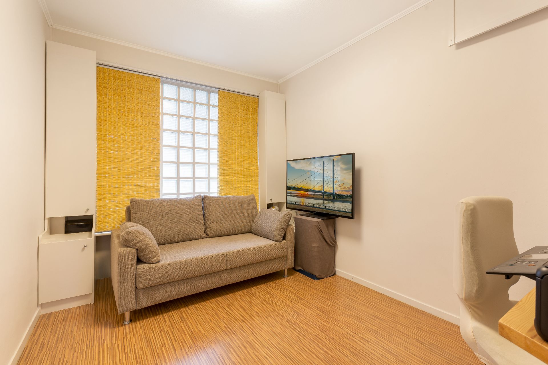 Kleines Wohnzimmer mit grauem Sofa, Fernseher, gelben Rollos und Holzfußboden.